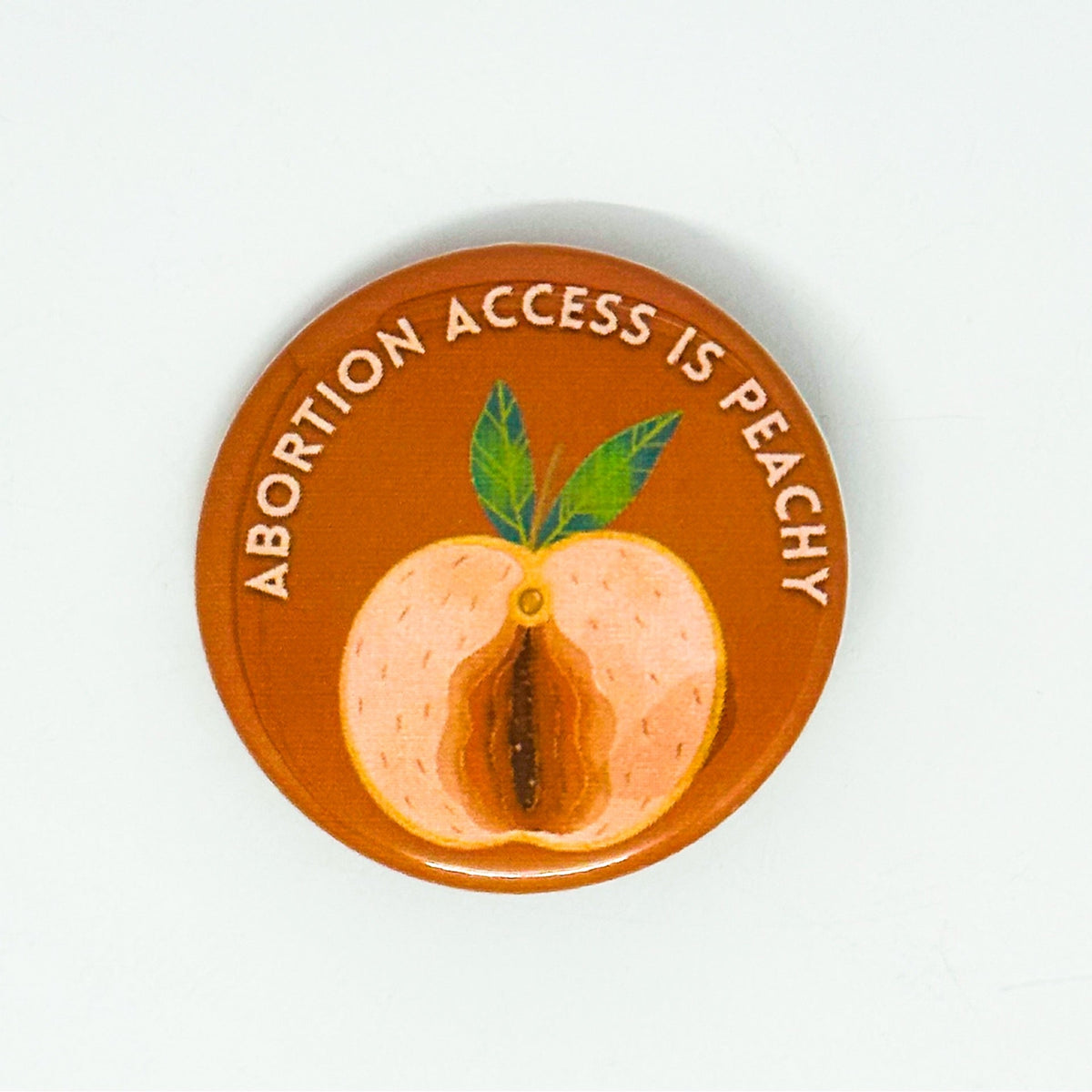 A round pin badge with a warm orange-brown background featuring a painted peach illustration and the text 'ABORTION ACCESS IS PEACHY' in white around the top edge.