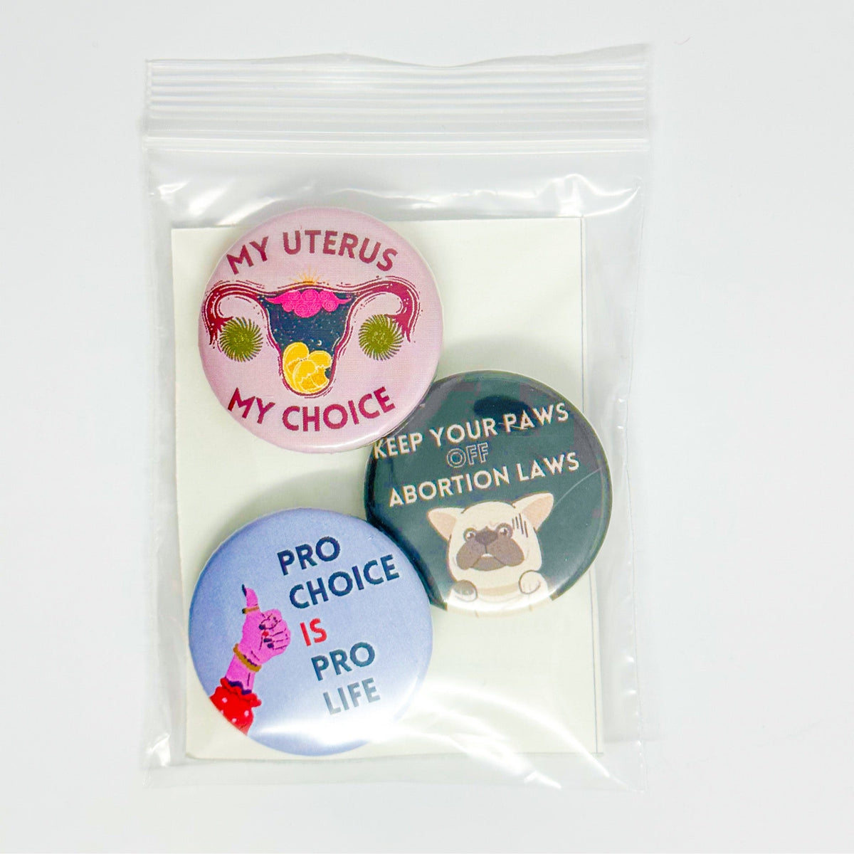 Three pro-choice themed button badges in a clear plastic bag with a white backing card. The badges feature illustrations and slogans including 'MY UTERUS MY CHOICE', 'KEEP YOUR PAWS OFF ABORTION LAWS', and 'PRO CHOICE IS PRO LIFE'.
