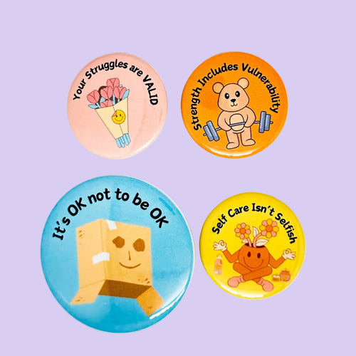 Four colorful round pin-back buttons: The buttons feature illustrated self-care themes: a pink button with flowers and the text 'Your Struggles are VALID,' an orange button with a bear lifting weights and 'Strength Includes Vulnerability,' a blue button with a cardboard box face and 'It's OK not to be OK,' and a yellow button with flowers and 'Self Care Isn't Selfish.