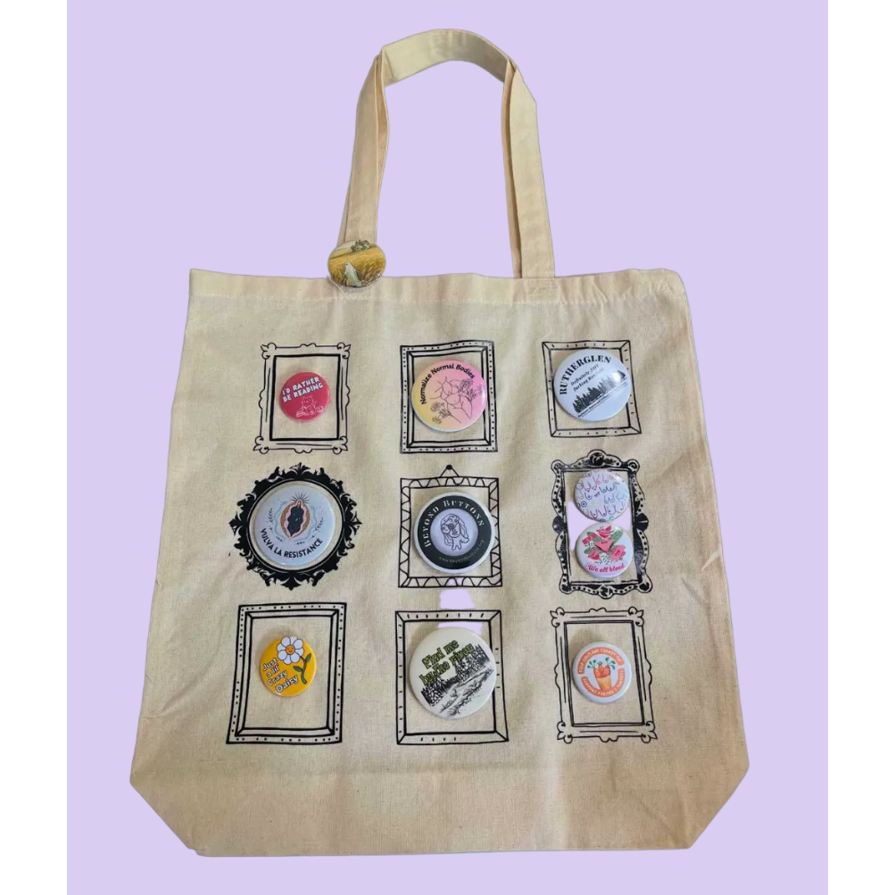 A beige canvas tote bag with nine decorative button frames attached to the front, set against a solid light blue background. 