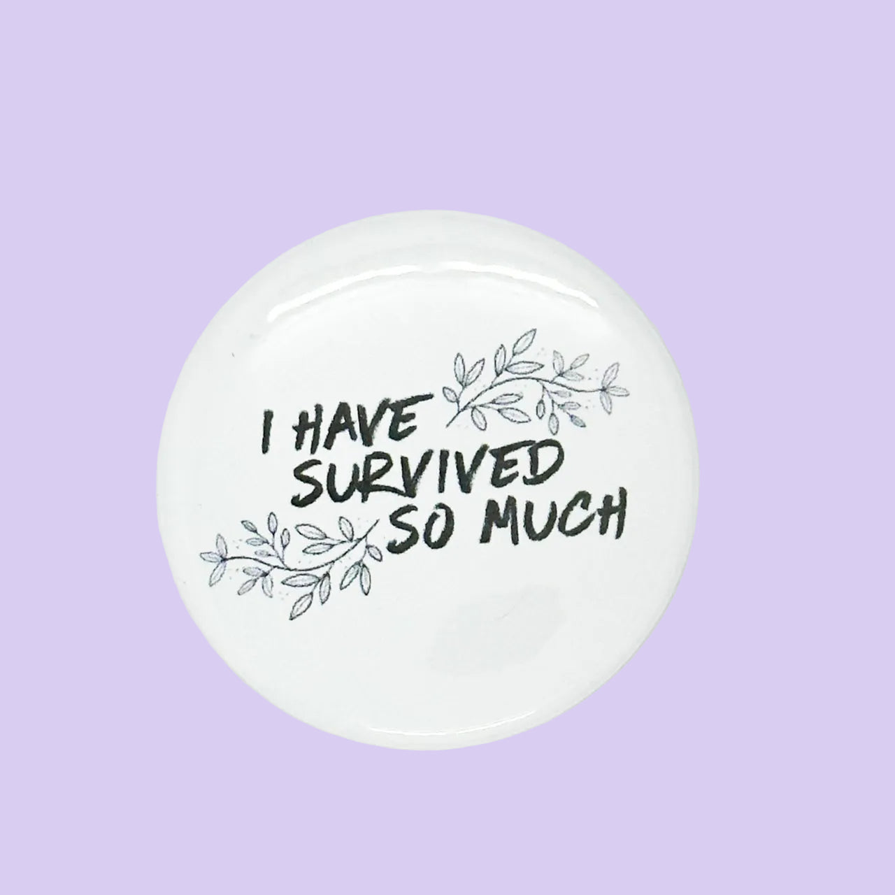 A round white button badge with the text 'I HAVE SURVIVED SO MUCH' in black handwritten-style lettering, decorated with delicate gray leaf illustrations, shown on a light purple background.