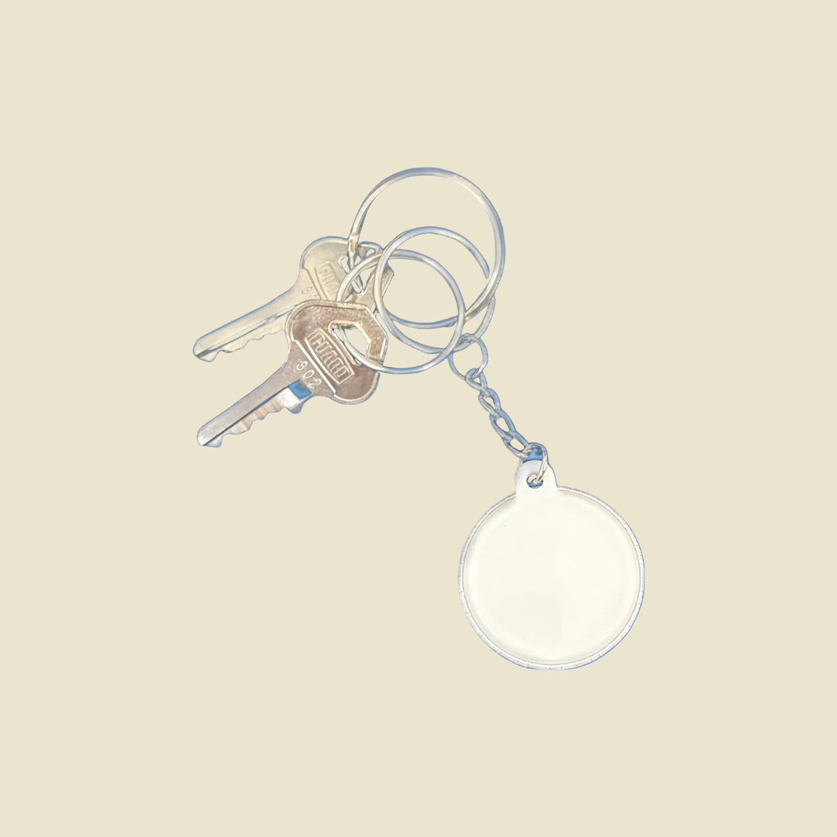 A silver metal keyring with two keys and a blank round keychain, shown against a light yellow background.