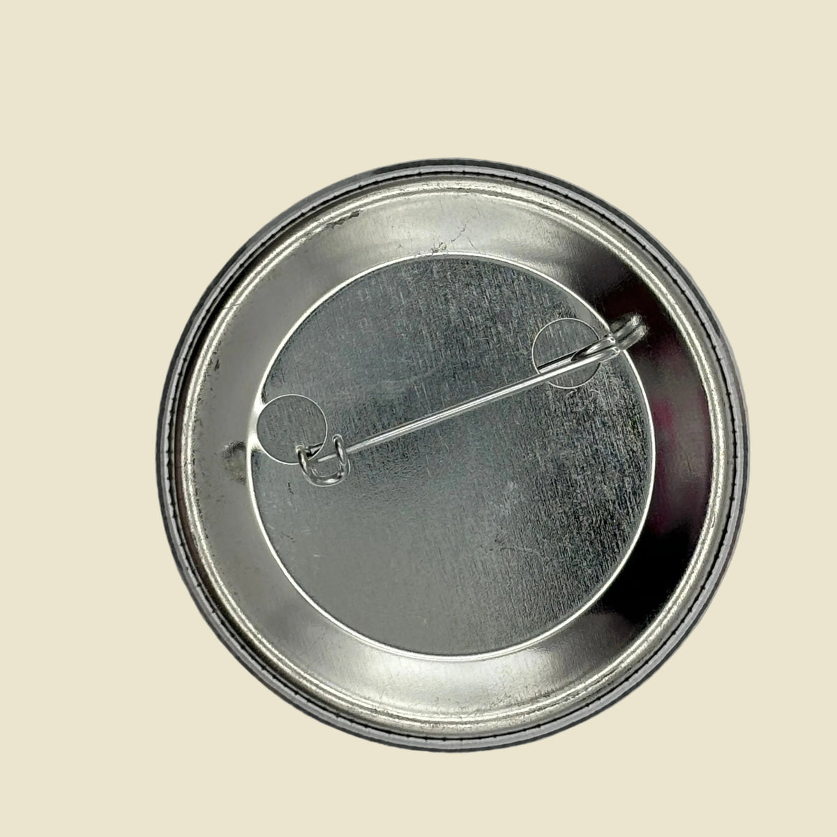 The back of a round metal pinback button badge, showing the silver-toned metal base and safety pin clasp mechanism on a light yellow background.